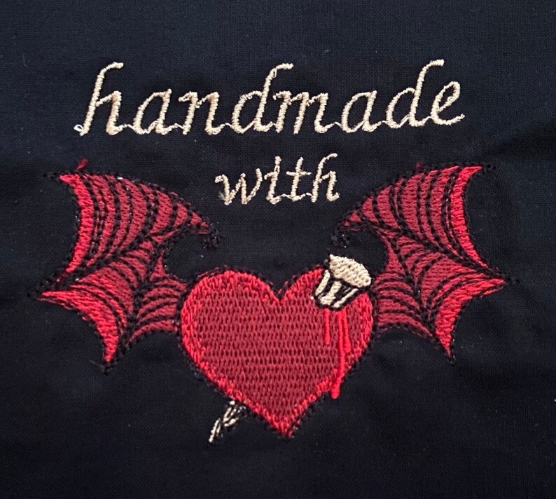 May include: Embroidered design with the text "handmade with" above a red heart with wings. The heart has a small white object on top and a red drip coming from it.