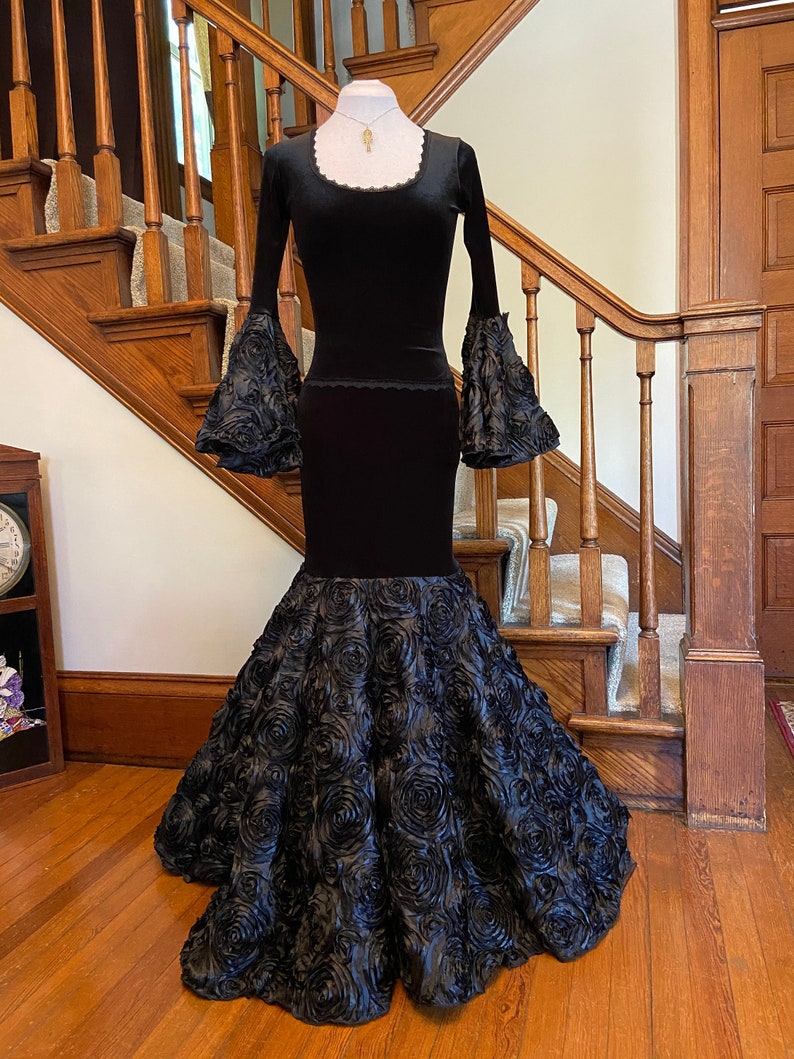 May include: A black velvet gown with long sleeves and a fitted bodice. The skirt is made of black fabric with a 3D rose design.
