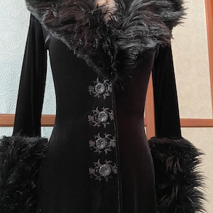 May include: A long black velvet coat with a faux fur collar and cuffs. The coat has a V-neckline and is fastened with buttons. The buttons are decorated with floral embroidery.