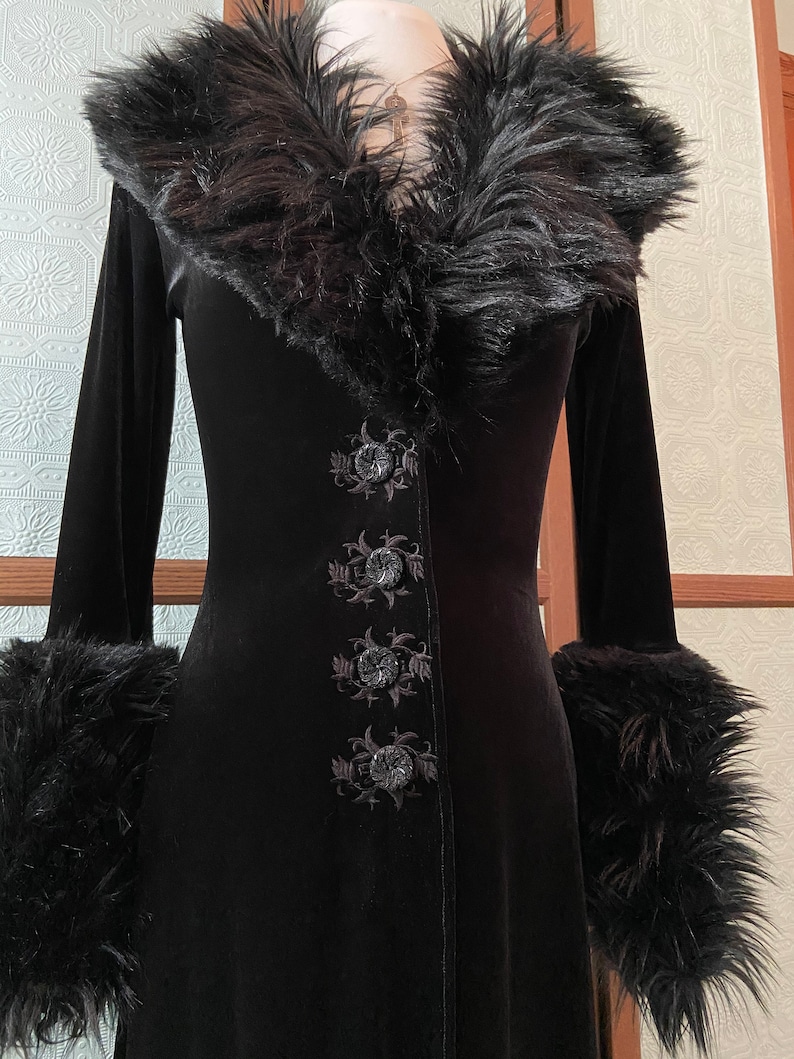 May include: A long black velvet coat with a faux fur collar and cuffs. The coat has a V-neckline and is fastened with buttons. The buttons are decorated with floral embroidery.