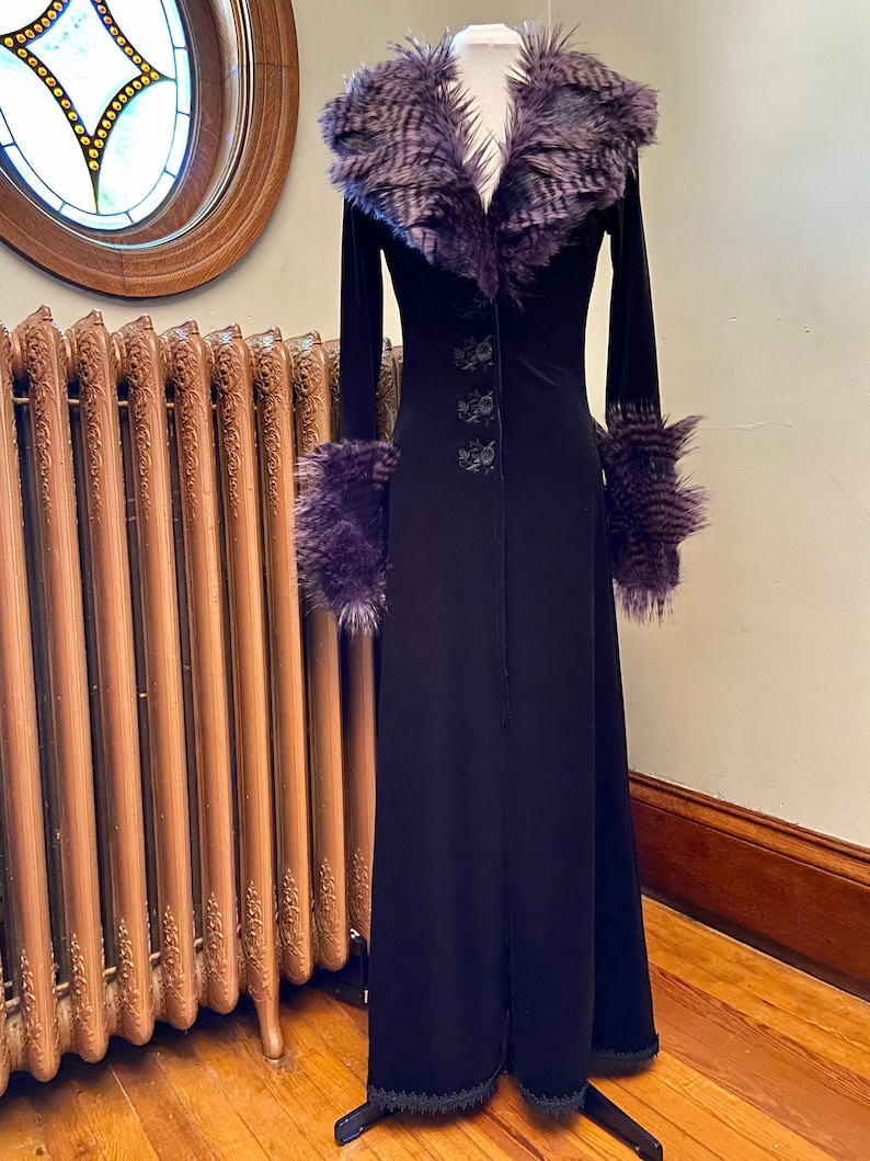 May include: A long black velvet coat with a purple faux fur collar and cuffs. The coat has a button closure and a lace trim at the hem.
