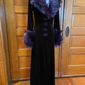 May include: A long black velvet coat with a purple faux fur collar and cuffs. The coat has a button closure and a lace trim at the hem.