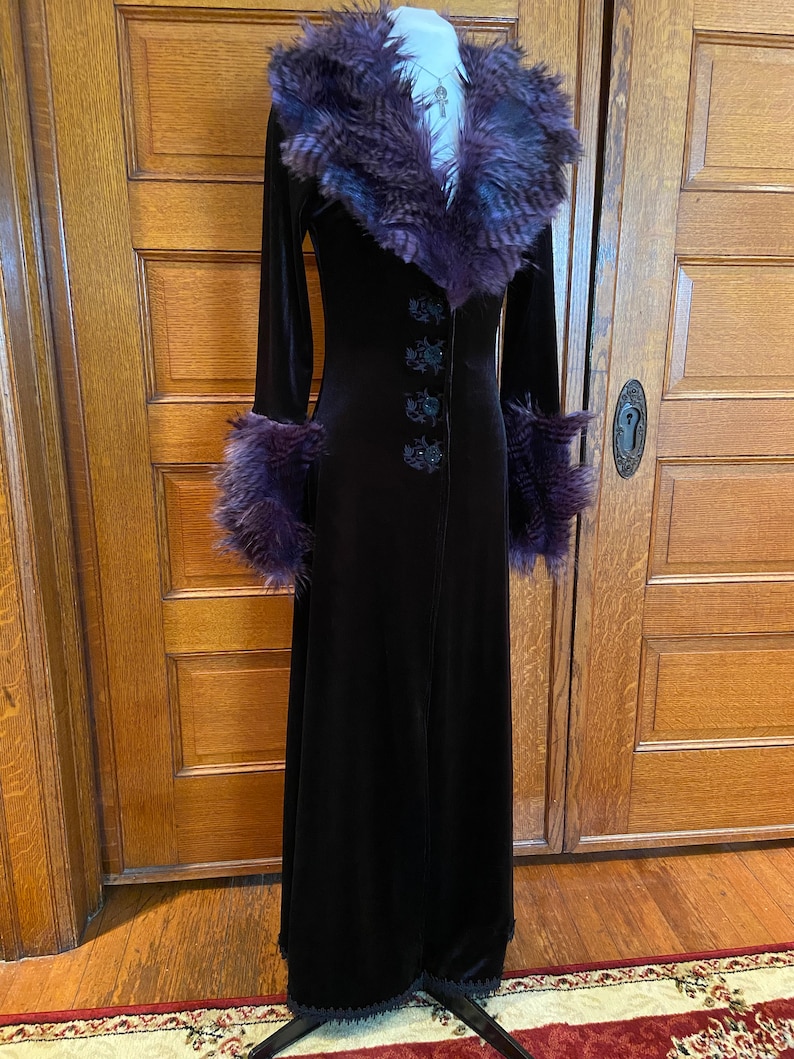 May include: A long black velvet coat with a purple faux fur collar and cuffs. The coat has a button closure and a lace trim at the hem.