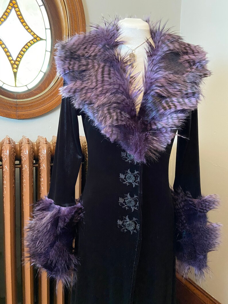 May include: A long black velvet coat with a purple faux fur collar and cuffs. The coat has a button closure and floral embroidery.