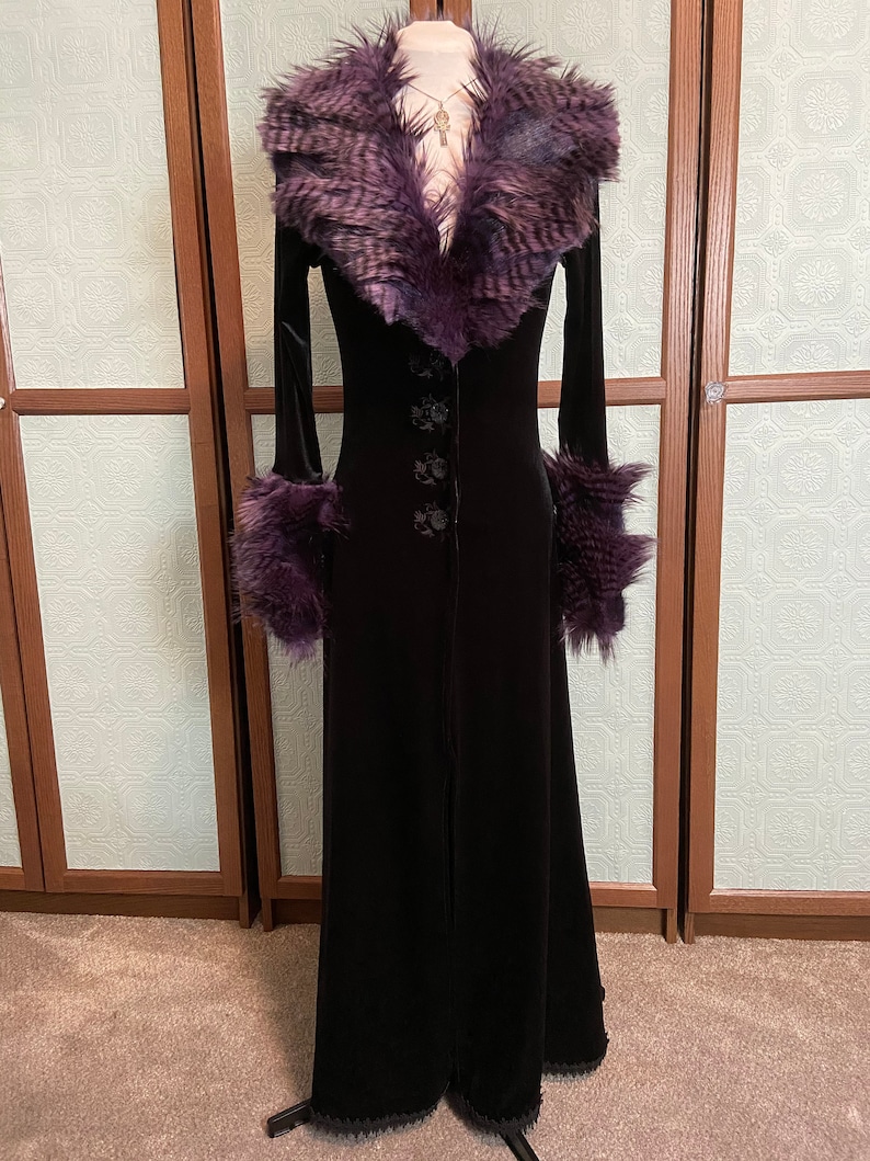 May include: A long black velvet coat with a purple faux fur collar and cuffs. The coat has a button closure and a fitted silhouette.