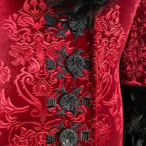 Op de afbeelding: Een rood fluwelen jasje met een zwarte bontkraag en zwarte geborduurde bloemdetails. Het jasje heeft een damastpatroon.