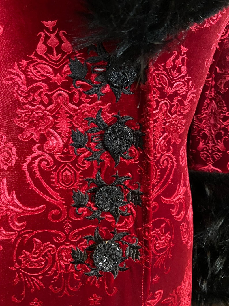 Op de afbeelding: Een rood fluwelen jasje met een zwarte bontkraag en zwarte geborduurde bloemdetails. Het jasje heeft een damastpatroon.