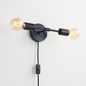 Puede incluir: Aplique de pared negro de dos bombillas con base redonda y brazo horizontal. El aplique tiene dos bombillas expuestas con un brillo amarillo cálido. Un cable negro con un interruptor de encendido/apagado cuelga debajo. Montado en una pared blanca.