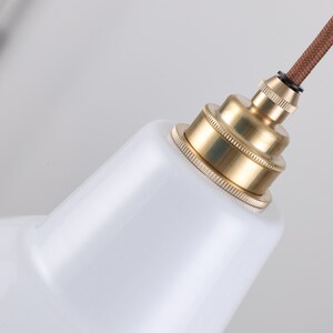 May include: Close-up of a white, cone-shaped pendant light fixture with a brass-colored socket and a brown fabric-covered cord. The fixture has a glossy finish and a vintage industrial design.