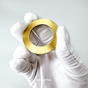 May include: A circular, gold-colored metal object with a hole in the center. The object has a brushed texture and is held by a gloved hand. The word "BRIGHTTIA" is visible at the bottom of the image.