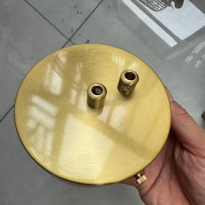 Puede incluir: Un dosel de techo redondo, color latón, con dos fijaciones cilíndricas y un pequeño tornillo. El dosel tiene un acabado cepillado y está diseñado para una lámpara. El diseño es simple y moderno.