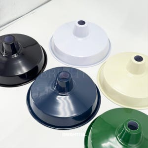 May include: Overhead view of five metal lamp shades in various colors: black, white, navy blue, cream, and green. Each shade has a rounded bowl shape with a central cylindrical opening. The word "BRIGHT" is partially visible on two shades.