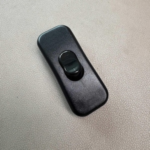 以下が含まれることがあります： 丸みを帯びたトグルボタンが付いた、黒色の長方形のオン/オフスイッチ。 スイッチは滑らかで光沢のある素材でできており、電気機器用に設計されています。 スイッチはニュートラルな色調の背景に表示されています。