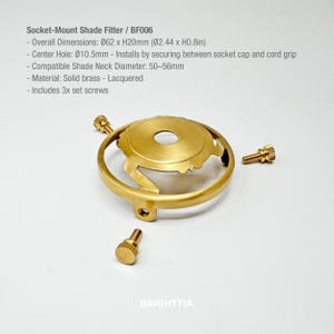 May include: A brass socket-mount shade fitter with three set screws. The fitter has overall dimensions of 2.44 inches in diameter and 0.8 inches in height. The center hole is 0.41 inches. The text on the image reads "Socket-Mount Shade Fitter / BF006".