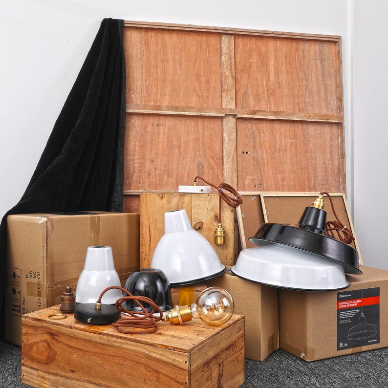 May include: A collection of industrial-style pendant lights in white and black, with brown cords and brass fixtures. The lights are displayed on wooden crates and cardboard boxes, with a large wooden panel in the background.