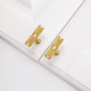 May include: Two gold-toned cabinet knobs with a textured, cylindrical design. The knobs are mounted on a white surface.
