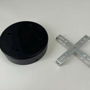 May include: A circular black metal ceiling canopy featuring four pre-drilled holes on the top surface and a side screw hole, accompanied by a separate silver-coloured metal cross-shaped mounting bracket with slotted openings for installation hardware.