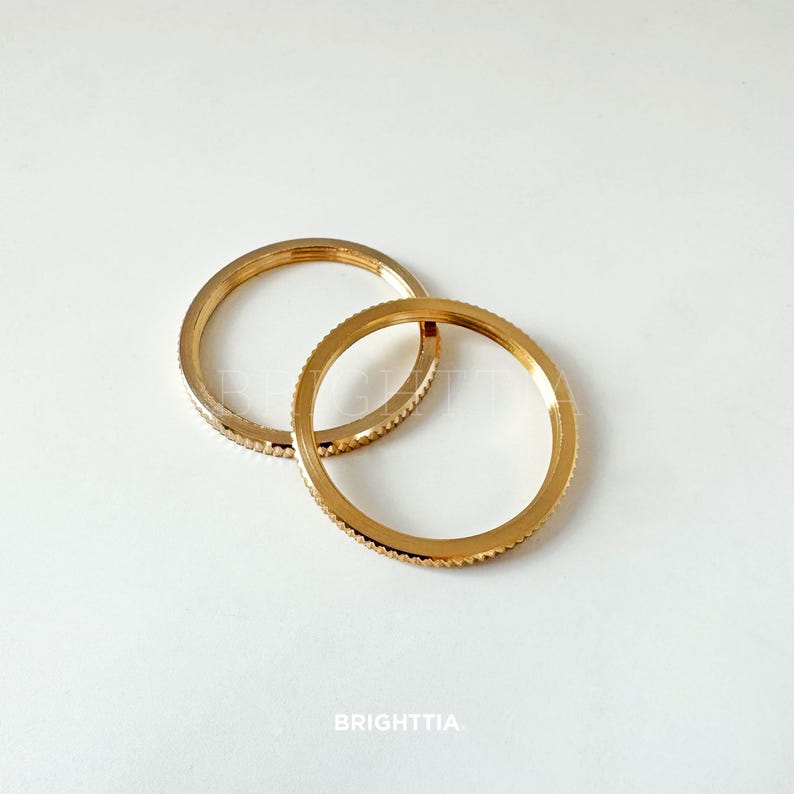Peut inclure: Deux anneaux m&eacute;talliques dor&eacute;s avec un motif textur&eacute; et stri&eacute;. Les anneaux sont circulaires et semblent &ecirc;tre faits d'un mat&eacute;riau m&eacute;tallique brillant. Le mot "BRIGHTTIA" est imprim&eacute; en bas de l'image.