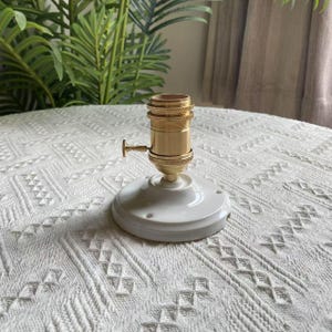 Peut inclure: Un support de lampe en laiton avec une base en c&eacute;ramique blanche. Le support a une forme cylindrique avec un petit interrupteur. La base est ronde avec des trous de montage. Le fond pr&eacute;sente de la verdure et une surface blanche &agrave; motifs.