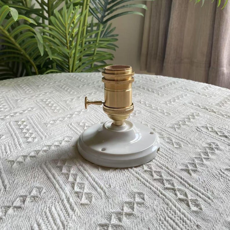 Peut inclure: Un support de lampe en laiton avec une base en c&eacute;ramique blanche. Le support a une forme cylindrique avec un petit interrupteur. La base est ronde avec des trous de montage. Le fond pr&eacute;sente de la verdure et une surface blanche &agrave; motifs.