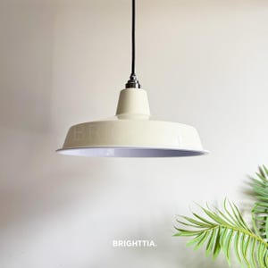Könnte beinhalten: Cremefarbene Pendelleuchte mit breitem, abgerundetem Schirm und weißem Innenraum. Die Leuchte hängt an einem schwarzen Kabel und trägt das Wort "BRIGHT" auf dem Schirm. Der Markenname "BRIGHTTIA" ist unten aufgedruckt.
