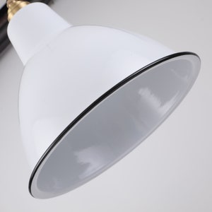 May include: Close-up of a white desk lamp with a black trim around the shade. The lamp has a brass-colored joint and a black arm. The interior of the shade is white.