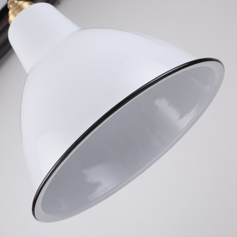 May include: Close-up of a white desk lamp with a black trim around the shade. The lamp has a brass-colored joint and a black arm. The interior of the shade is white.