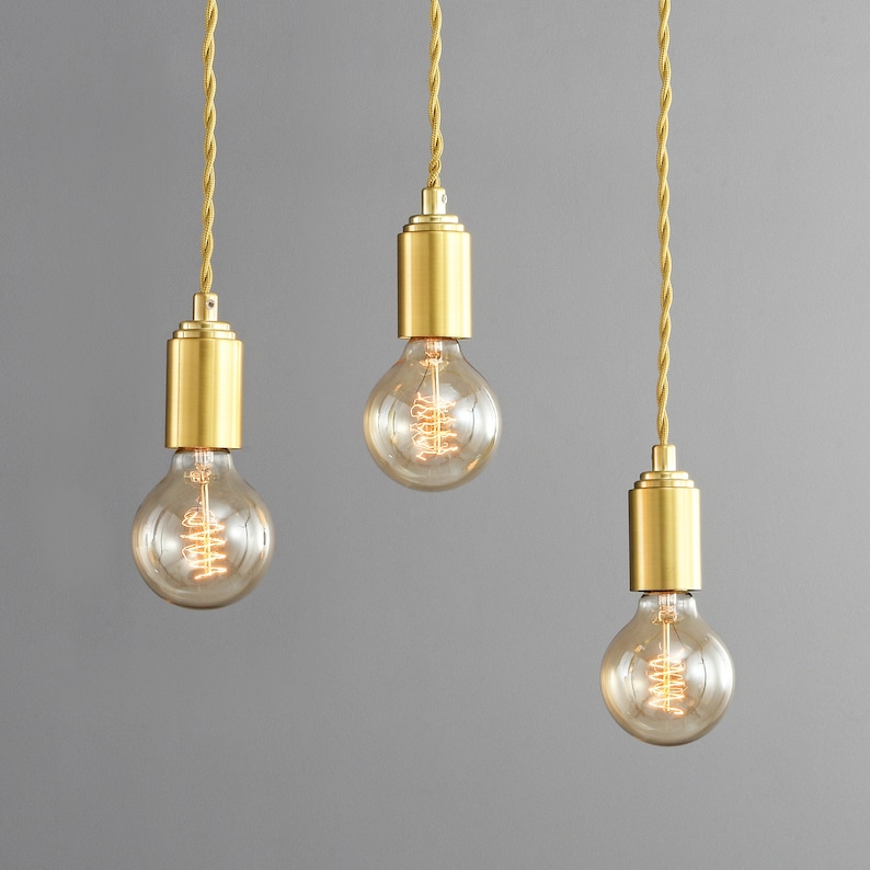 Three STEP UP brushed gold hanging pendant lights with globe style Edison light bulbs hung in a row. Ceiling mounted on a gray background.