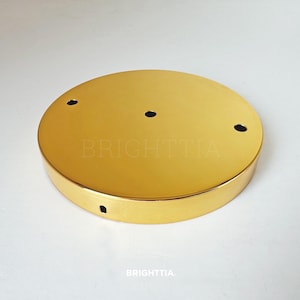 Puede incluir: Una roseta de techo redonda, de color dorado, con cuatro agujeros espaciados uniformemente. El producto tiene una superficie brillante y reflectante. La palabra "BRIGHTTIA" está impresa en la superficie.
