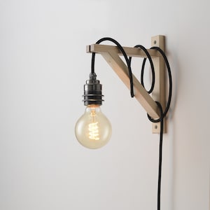 Op de afbeelding: Een houten wandlamp met een zwart snoer en een gloeilamp. De lamp is gemaakt van natuurlijk hout en heeft een eenvoudig, modern ontwerp.