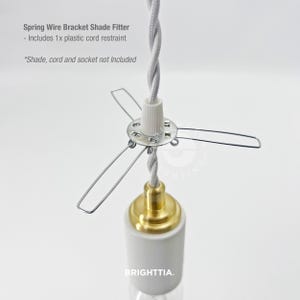 May include: A spring wire bracket shade fitter with a white plastic cord restraint. The fitter has a silver metal frame with a white and gray twisted cord. The bottom features a gold-colored socket and a white ceramic base. Text on the image reads "BRIGHTTIA."