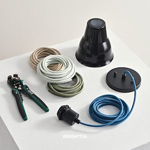 May include: An assortment of electrical components for a pendant light fixture. Includes a black lamp shade, a black ceiling canopy, a black light socket, and coiled fabric-covered wires in beige, light green, and olive green. A blue fabric-covered wire and a wire stripper are also present.