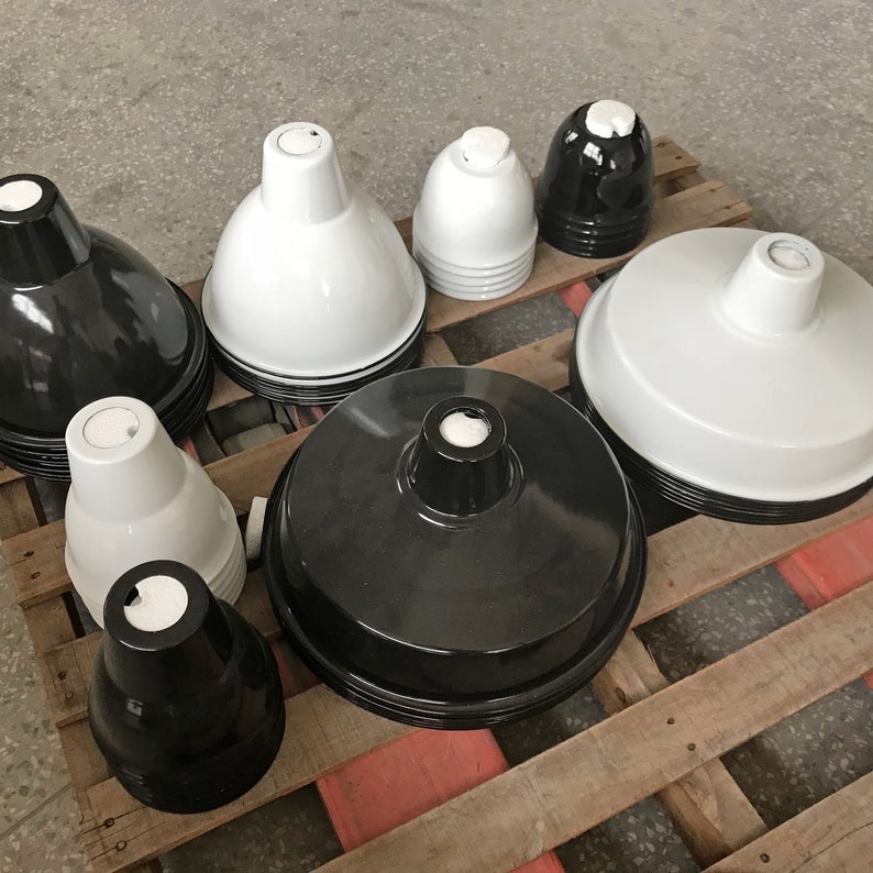 May include: A collection of glossy, industrial-style pendant light shades in various sizes and shapes. The shades are primarily black and white, with a few gray ones. They are arranged on a wooden pallet, showcasing their design and finish.