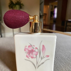 May include: A vintage perfume bottle with a white rectangular body and a maroon atomizer. The bottle features a pink floral design. The atomizer is gold-toned.