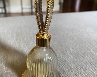 Puede incluir: Una botella de vidrio transparente con forma de campana con una tapa de metal dorado y un asa decorativa. La botella tiene un diseño acanalado.