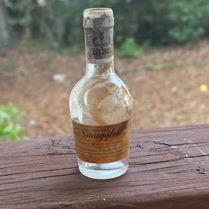 Puede incluir: Una botella de vidrio en miniatura de Old Smuggler Blended Scotch Whisky. La botella tiene un cuerpo de vidrio transparente y una etiqueta de papel marrón con letras doradas. La etiqueta dice "Old Smuggler Blended Scotch Whisky" e incluye el año "1835".