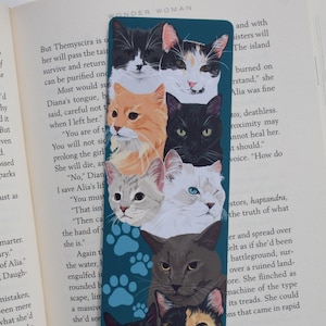 Puede incluir: Un marcapáginas azul verdoso con una ilustración colorida de seis gatos. Los gatos están apilados, con sus cabezas y hombros visibles. Los gatos son todos de diferentes colores y patrones, incluyendo negro, blanco, naranja y calico. El marcapáginas tiene un patrón de huellas de patas en el fondo.