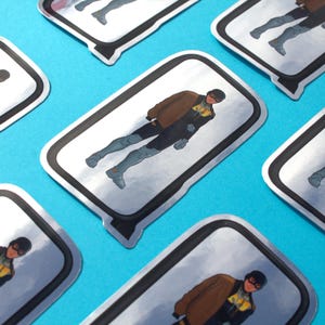 Op de afbeelding: Meerdere stickers met een superheld in een bruine jas, zwart pak en grijze laarzen. De stickers hebben een zilveren rand en zijn geplaatst op een helderblauwe achtergrond. De superheld draagt een zwart masker met gele accenten.