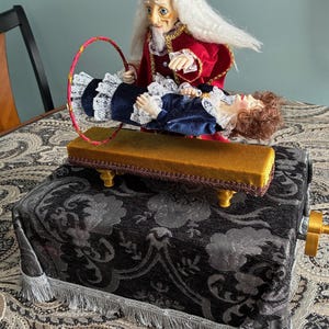 Può includere: Un giocattolo vintage di illusionismo da mago con due bambole dettagliate. Una, vestita con cappotto e cappello rossi, tiene un cerchio sopra una bambola con un vestito blu. Le bambole sono su una piattaforma dorata sopra una scatola fantasia con una manovella.