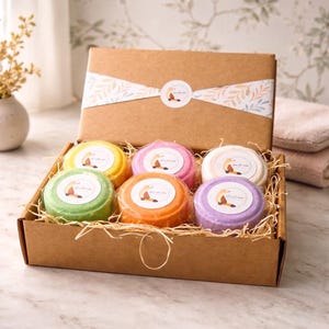 Puede incluir: Una caja de regalo de cartón marrón que contiene seis bombas de baño redondas y coloridas en amarillo, rosa, blanco, verde, naranja y morado. Cada bomba está envuelta individualmente en plástico transparente con una pegatina de un zorro.