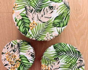 May include: Three round fabric bowl covers with a tropical leaf and floral pattern. The covers feature green palm leaves, light pink leaves, and yellow and pink flowers on a white background. The covers are different sizes.