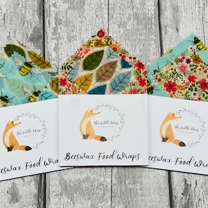 May include: Three beeswax food wraps with different patterns. The wraps are folded and displayed on a white wooden surface. Each wrap has a label with the text "Beeswax Food Wraps" and a logo of a fox in a wreath. The patterns include bees and flowers on a light blue background, leaves on a cream background, and flowers on a light blue background.