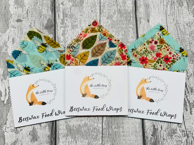 May include: Three beeswax food wraps with different patterns. The wraps are folded and displayed on a white wooden surface. Each wrap has a label with the text "Beeswax Food Wraps" and a logo of a fox in a wreath. The patterns include bees and flowers on a light blue background, leaves on a cream background, and flowers on a light blue background.