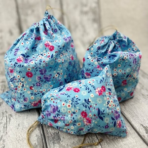 Könnte beinhalten: Drei blaue Stoffbeutel mit Kordelzug und einem Blumenmuster in Rosa-, Lila- und Weißtönen. Die Beutel sind aus einem weichen, leichten Stoff gefertigt und haben ein schlichtes, elegantes Design.