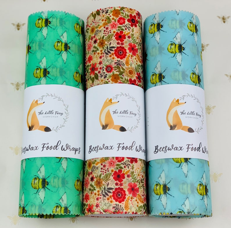 May include: Three beeswax food wraps with different designs. The first wrap has a green background with yellow bees, the second wrap has a floral pattern with red, yellow, and orange flowers, and the third wrap has a light blue background with yellow bees. Each wrap has a label that says "The Little Foxy Workshop" and "Beeswax Food Wraps".