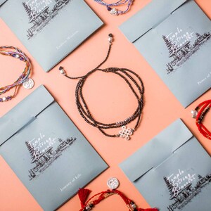 May include: A selection of Temple String bracelets displayed on a peach surface. The bracelets feature diverse colours, such as black, red, and purple, and include decorative charms. Light blue envelopes with the text "Temple String Bracelet" are arranged around the bracelets.