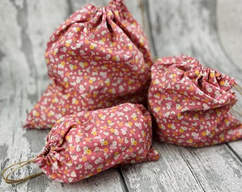 May include: Three pink drawstring bags with a floral pattern. The bags are made of fabric and have a white background with pink, yellow, and green flowers.