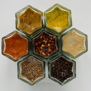 Op de afbeelding: Zes zeshoekige glazen potten met metalen deksels die verschillende kruiden bevatten. De potten zijn in een honingraatpatroon gerangschikt. De kruiden omvatten rode pepervlokken, zwarte peperkorrels, gedroogde kruiden en gemalen kruiden.