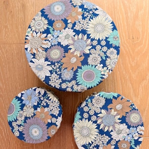 Puede incluir: Tres cubiertas redondas de tela para cuencos con un estampado floral. Las cubiertas presentan un fondo azul marino con un patrón repetido de flores blancas, celeste, naranjas y moradas. Las cubiertas son de diferentes tamaños.
