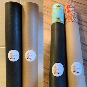 May include: Two cardboard tubes with black and brown paper wrapping. Each tube has a sticker with a fox illustration and the text "The Little Fox" on it. The tubes are filled with two different patterned beeswax food wraps. One wrap has a blue background with bees and the other has a floral pattern.